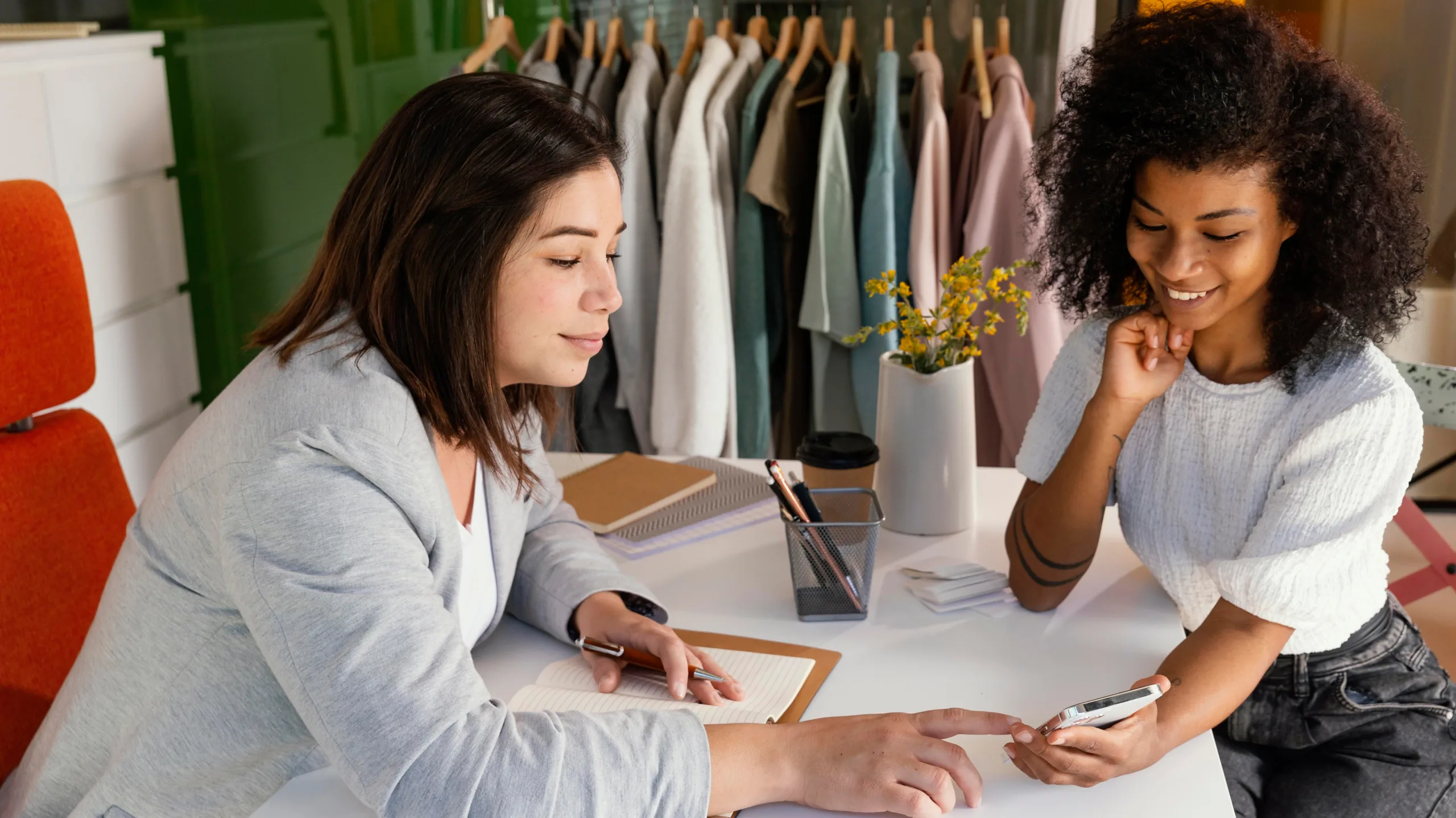 Two women discussing clothing options during a tailoring consultation with garments hanging in the background